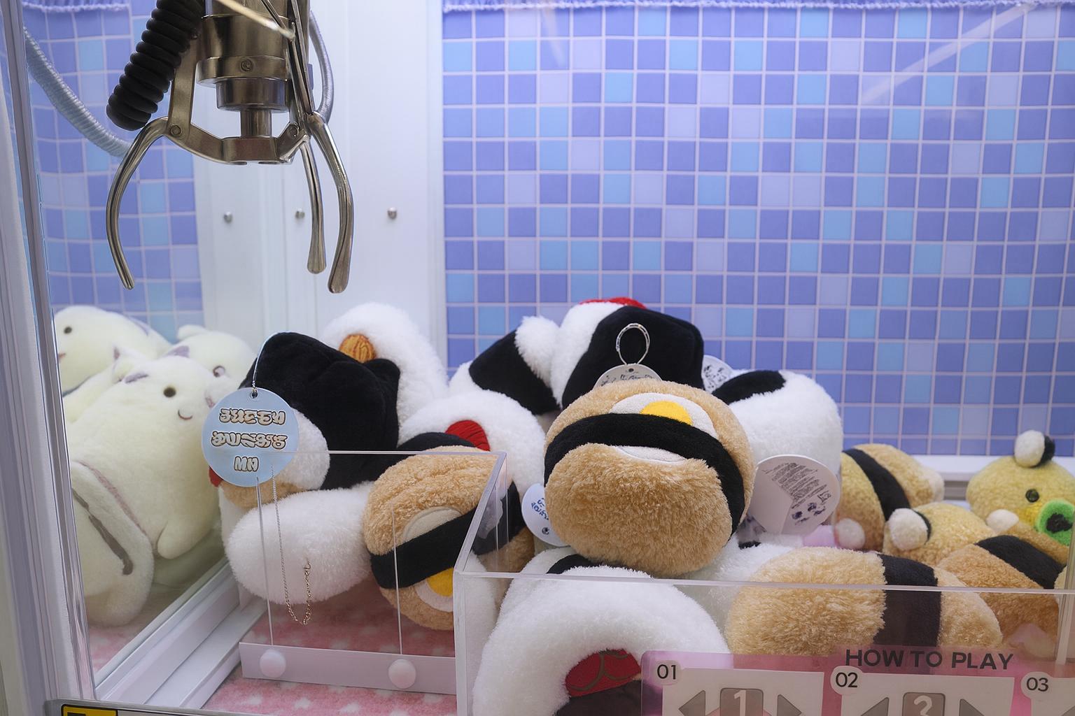 ローソン店舗に設置されたクレーンゲーム機の内部。青いタイル模様の背景に、おにぎり型やぬいぐるみが並び、金属のアームが上から下がっている様子